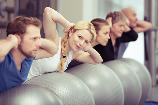 Eine Gruppe von fünf Personen übt auf Gymnastikbällen, einige halten die Hände hinter dem Kopf.