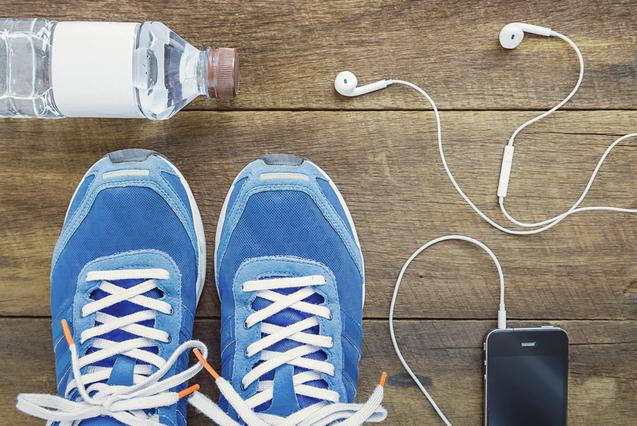 Blick von oben auf blaue Sportschuhe, Wasserflasche, Kopfhörer und ein Smartphone auf einer Holzoberfläche.