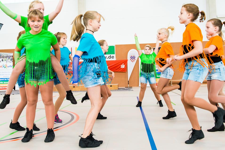 Gruppe von Tänzerinnen in bunten Shirts und Shorts führt synchronisierte Bewegungen auf einer Sportfläche aus.