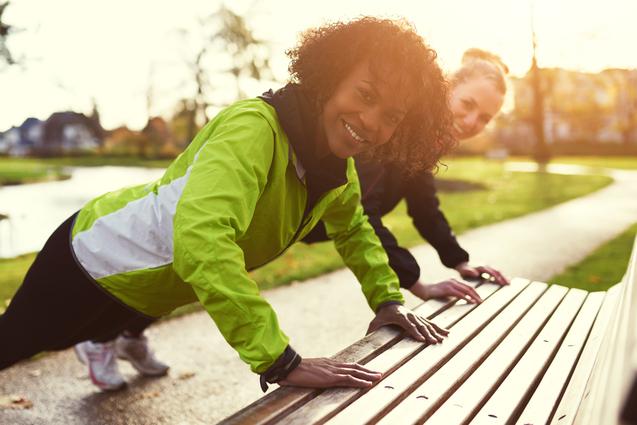 Zwei Frauen in Sportkleidung machen im Park Liegestütze auf einer Holzbank, die Sonne scheint.