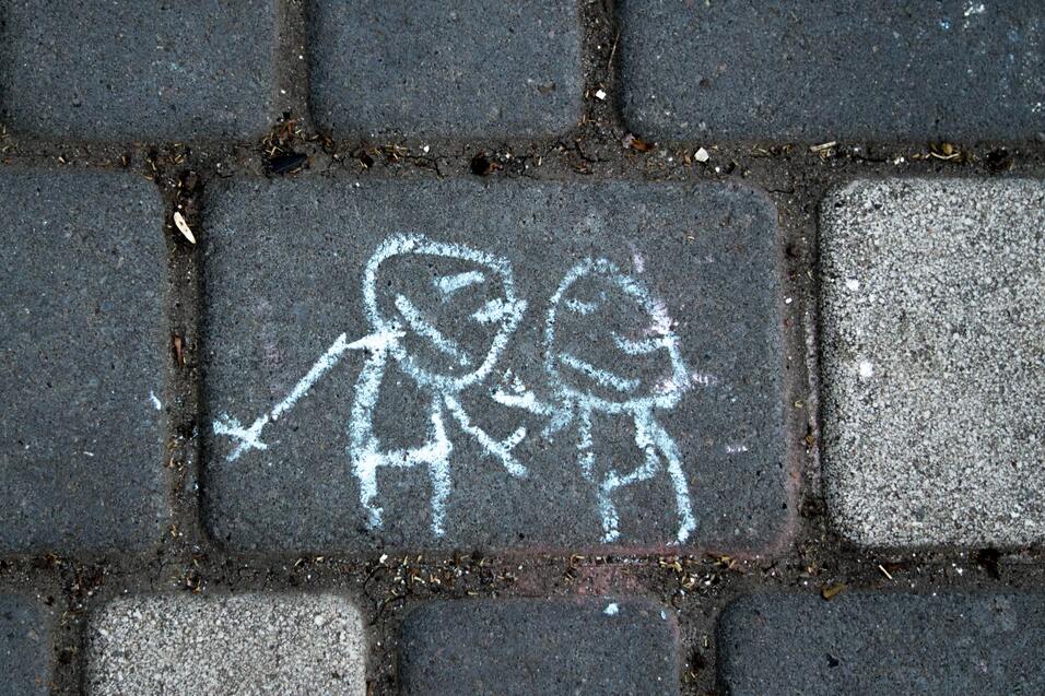 Kreidezeichnung von zwei Figuren, die sich an den H&auml;nden halten, auf einem grau gepflasterten Boden.