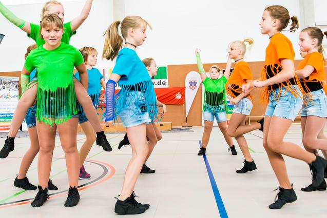 Gruppe von Tänzerinnen in bunten Shirts und Shorts führt synchronisierte Bewegungen auf einer Sportfläche aus.