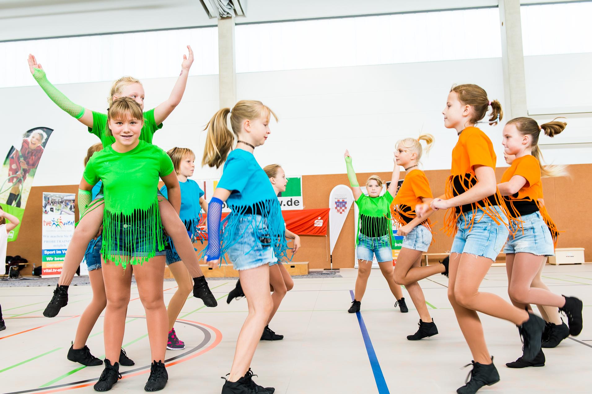 Gruppe von Tänzerinnen in bunten Shirts und Shorts führt synchronisierte Bewegungen auf einer Sportfläche aus.