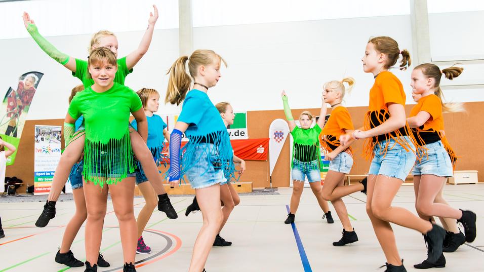Gruppe von Tänzerinnen in bunten Shirts und Shorts führt synchronisierte Bewegungen auf einer Sportfläche aus.