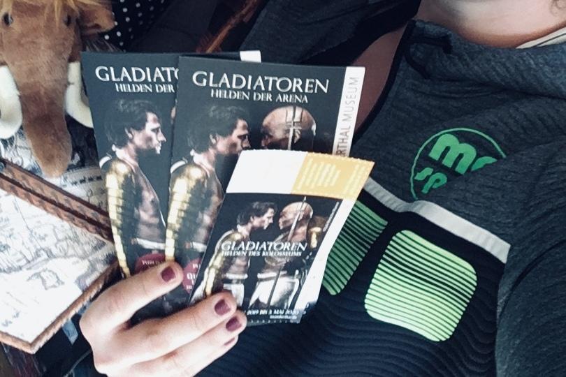 Zwei Frauen posieren mit Eintrittskarten f&uuml;r die Ausstellung "Gladiatoren" vor einer holzget&auml;felten Wand.