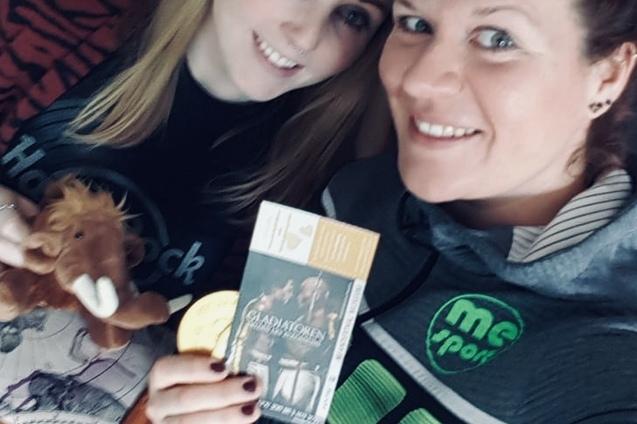 Zwei Frauen posieren nah beieinander und halten eine Eintrittskarte und ein Stofftier vor einem Bücherregal.