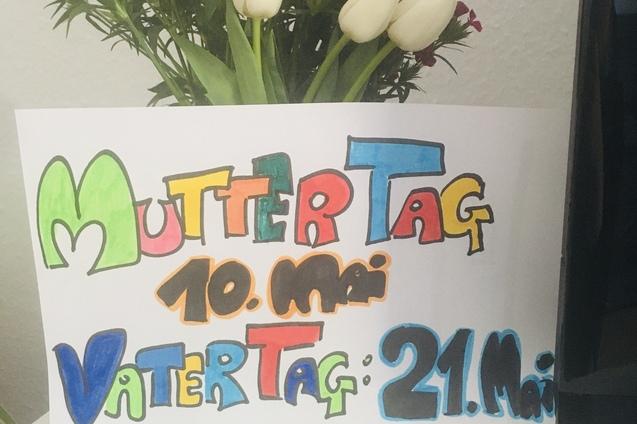 Bunt beschriftetes Papier mit „Muttertag 10. Mai“ und „Vattertag 21. Mai“ steht neben einer Blumenvase mit Tulpen.