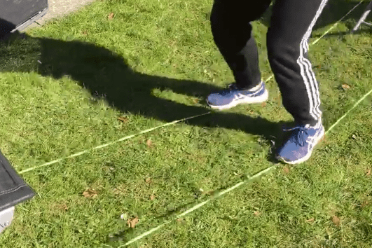 Person balanciert auf einer grünen Slackline, mit Schatten auf der grünen Wiese und einem Liegestuhl im Hintergrund.