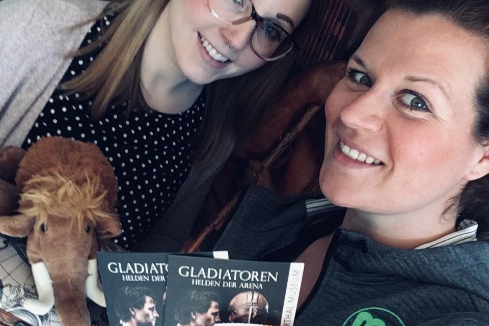 Zwei Frauen posieren mit Eintrittskarten für die Ausstellung "Gladiatoren" vor einer holzgetäfelten Wand.