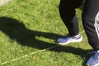 Person balanciert auf einer grünen Slackline, mit Schatten auf der grünen Wiese und einem Liegestuhl im Hintergrund.