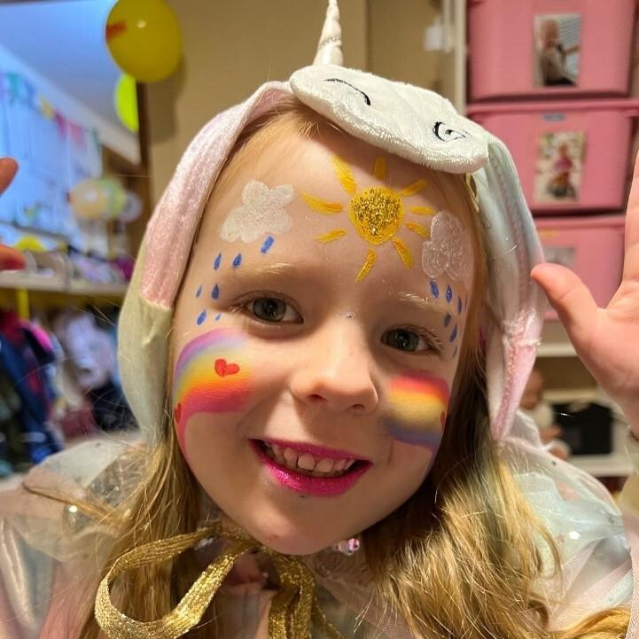 Mädchen mit Einhorn-Haube und buntem Gesichtsmalerei, lächelt fröhlich in einem feierlichen Raum.