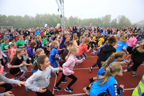 Viele Kinder in Sportkleidung laufen auf einer roten Laufbahn, umgeben von Zuschauern bei einem Sportevent.