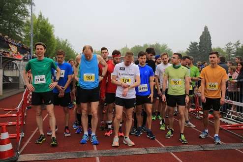 Gruppe von Laufteilnehmern in Sportkleidung, bereit zum Wettstart auf einer Laufbahn, Wettkampfnummern sichtbar.