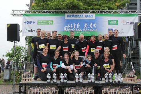 Gruppe von Laufteilnehmern in schwarzen T-Shirts posiert auf Podium mit Pokalen und Bannern beim Mettmanner Bachlauf.