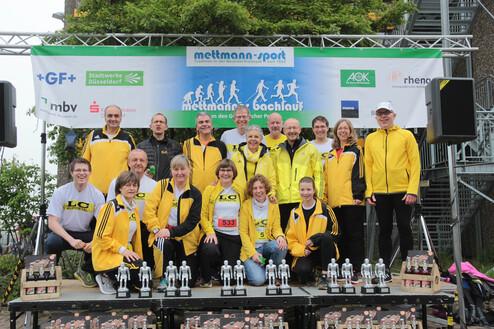 Gruppe von Läufern in gelben Outfits steht vor einer Bühne mit Pokalen, Banner und Sponsorenlogos beim Mettmanner Bachlauf.