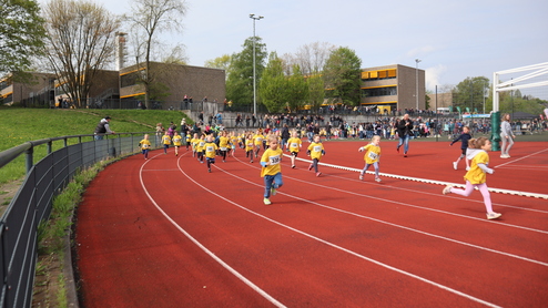 Eine Gruppe von Kindern in gelben T-Shirts läuft auf einer roten Laufbahn, umgeben von Zuschauern und Bäumen.
