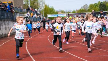 Gruppe von Kindern in Sportkleidung laufen auf einer roten Laufbahn während eines Wettkampfs. Zuschauer beobachten vom Rand.