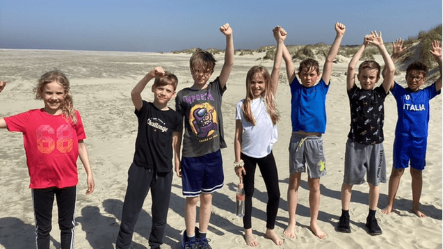 Sechs Kinder stehen am Strand, heben die Arme und schauen in die Kamera, umgeben von Sand und Dünen.