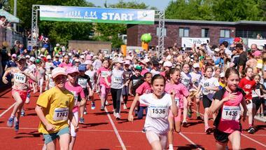 Kinder laufen auf einer roten Laufbahn bei einem Wettkampf, begleitet von begeisterten Zuschauern im Hintergrund.