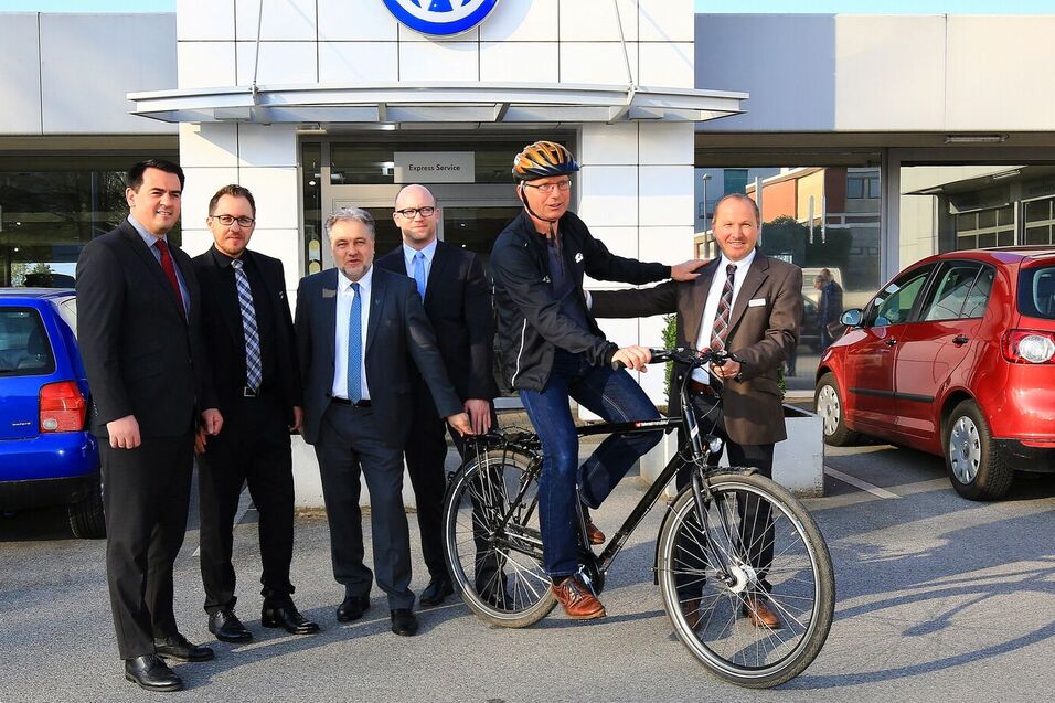 Sechsmal Männer posieren vor einem Volkswagen-Händler, ein Mann sitzt auf einem Fahrrad. Zwei Autos stehen im Hintergrund.