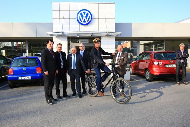 Sechsmal Männer posieren vor einem Volkswagen-Händler, ein Mann sitzt auf einem Fahrrad. Zwei Autos stehen im Hintergrund.