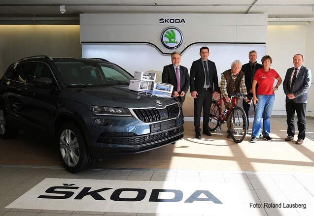 Personengruppe steht neben einem schwarzen Škoda-Auto, dazu ein rotes Fahrrad und einige Geschenkboxen in einem Autohaus.