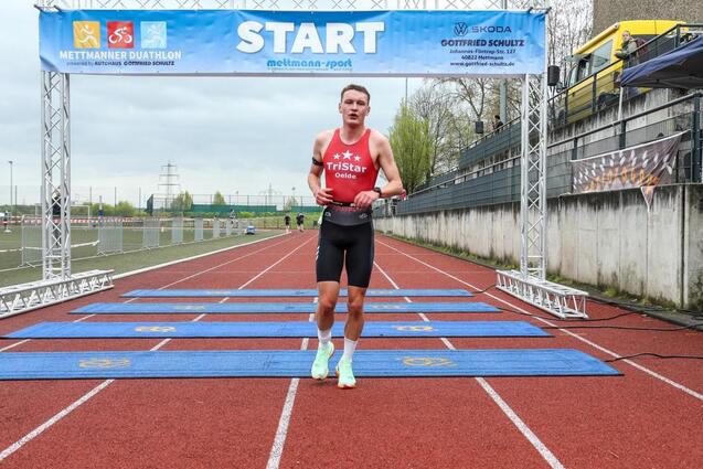 Teilnehmer beim Mettmanner Duathlon läuft über die Startlinie auf einem roten Sportplatz, umgeben von Zuschauern.