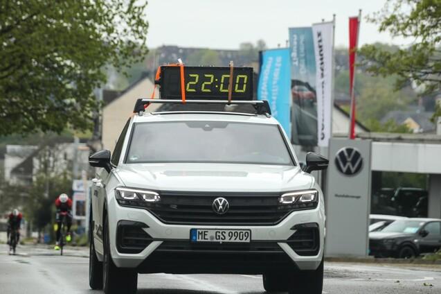 Weißer Volkswagen SUV mit digitaler Anzeige "22:00", während Radfahrer im Hintergrund eine Straße entlangfahren.