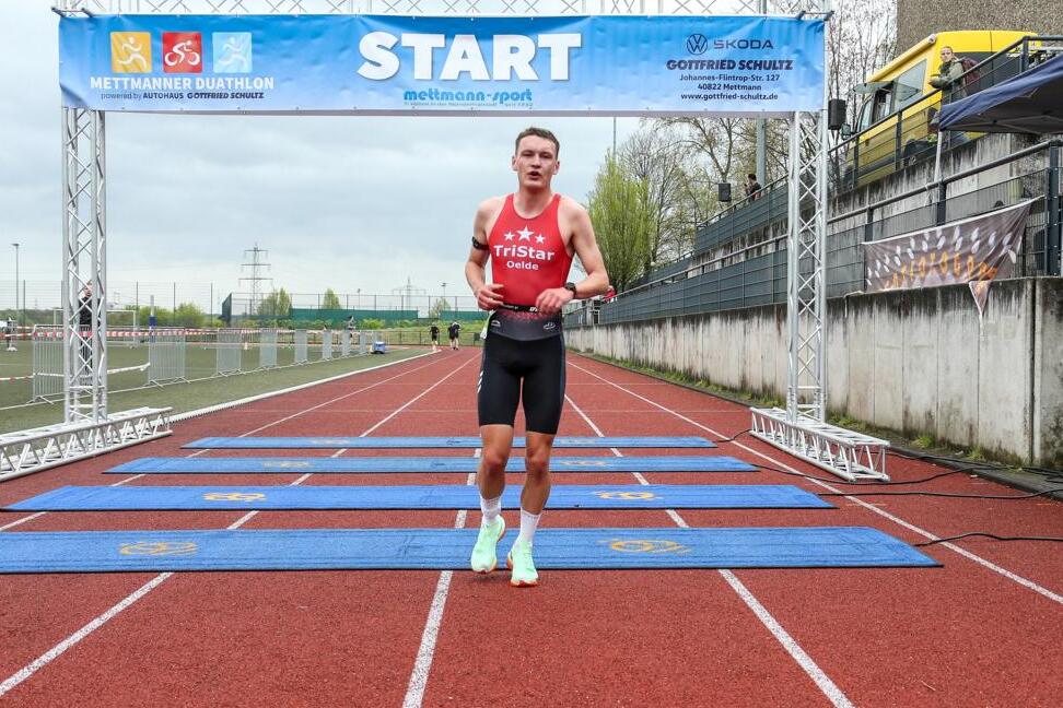 Teilnehmer beim Mettmanner Duathlon läuft über die Startlinie auf einem roten Sportplatz, umgeben von Zuschauern.