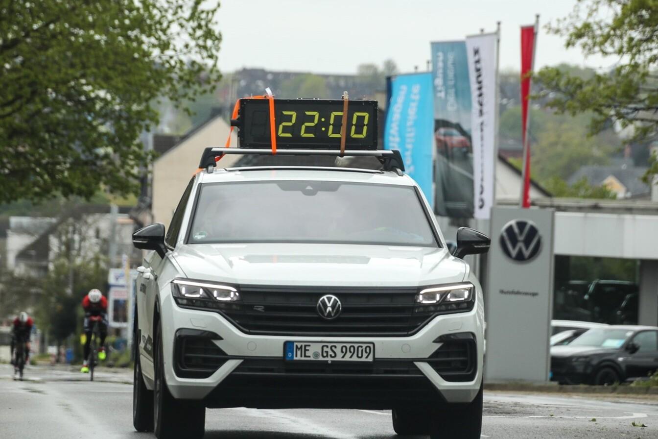 Weißer Volkswagen SUV mit digitaler Anzeige "22:00", während Radfahrer im Hintergrund eine Straße entlangfahren.