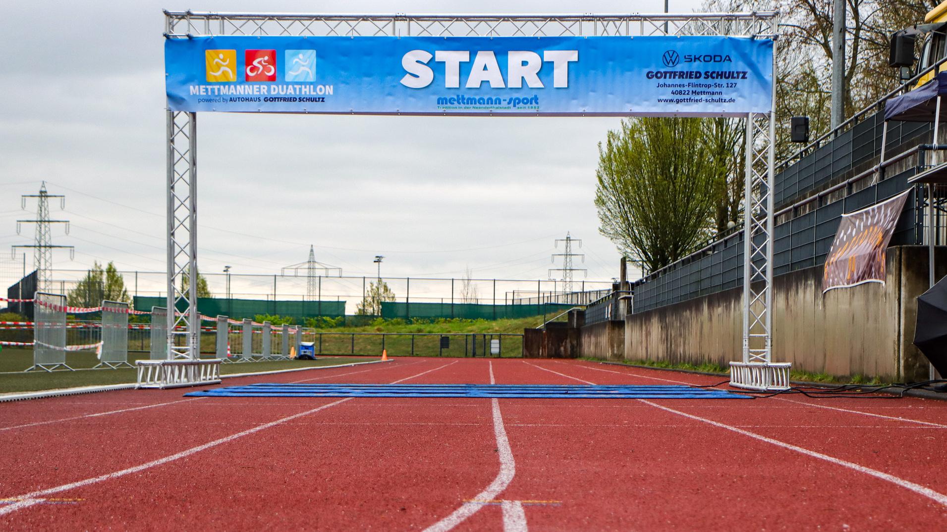 Startbogen über einer roten Laufbahn, mit Werbung für den Mettmann Duathlon und einem blauen Startbereich.