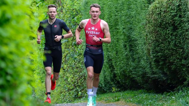 Zwei Triathleten laufen auf einem schmalen Weg entlang grüner Hecken in einer vom Gras gesäumten Landschaft.
