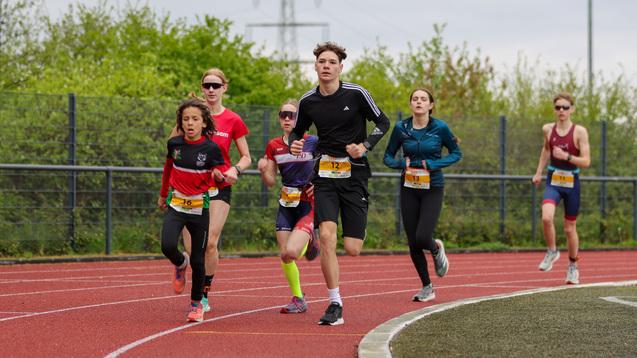 Sechs Jugendliche laufen auf einer roten Laufbahn, umgeben von grünem Nachwuchs und einem bewölkten Himmel.