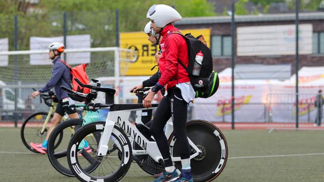 Zwei Sportler in Rennkleidung gehen mit ihren Fahrrädern auf einem grünen Sportplatz entlang.