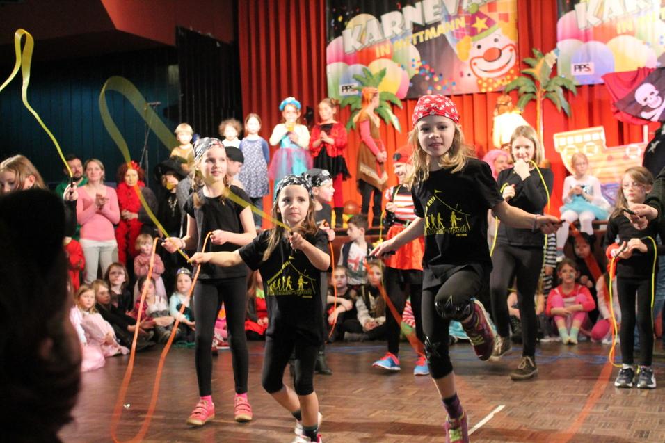 Kinder in Karnevalskostümen springen mit bunten Seilen auf einer Bühne vor einem festlichen Hintergrund.