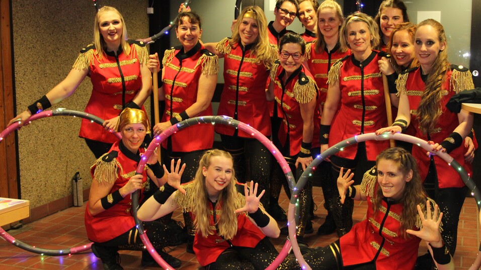 Gruppennahaufnahme von zwölf Frauen in roten Kostümen mit Hula-Hoops, die fröhlich in eine Kamera lächeln.