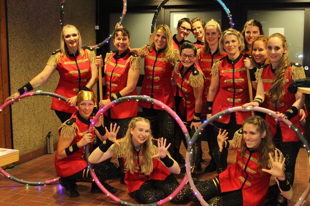 Gruppennahaufnahme von zw&ouml;lf Frauen in roten Kost&uuml;men mit Hula-Hoops, die fr&ouml;hlich in eine Kamera l&auml;cheln.