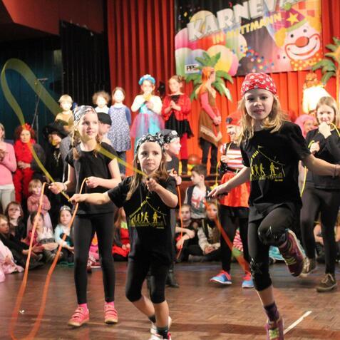 Kinder in Karnevalskostümen springen mit bunten Seilen auf einer Bühne vor einem festlichen Hintergrund.