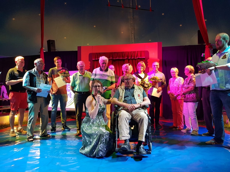 Vielseitige Gruppenfeier mit Medaillenverleih, wo eine Frau auf einem Podium spricht und mehrere Personen mit Blumen posieren.