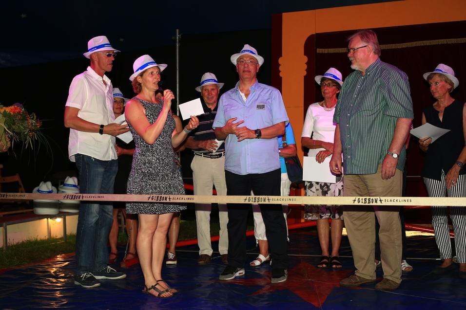 Gruppe von Personen in Hüten, die auf einer Bühne sprechen und applaudieren, umgeben von festlicher Dekoration.