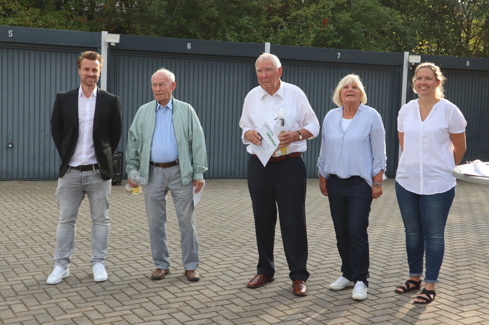Fünf Personen stehen lächelnd in einer Reihenfolge vor einer grauen Garage, einige halten Dokumente in der Hand.