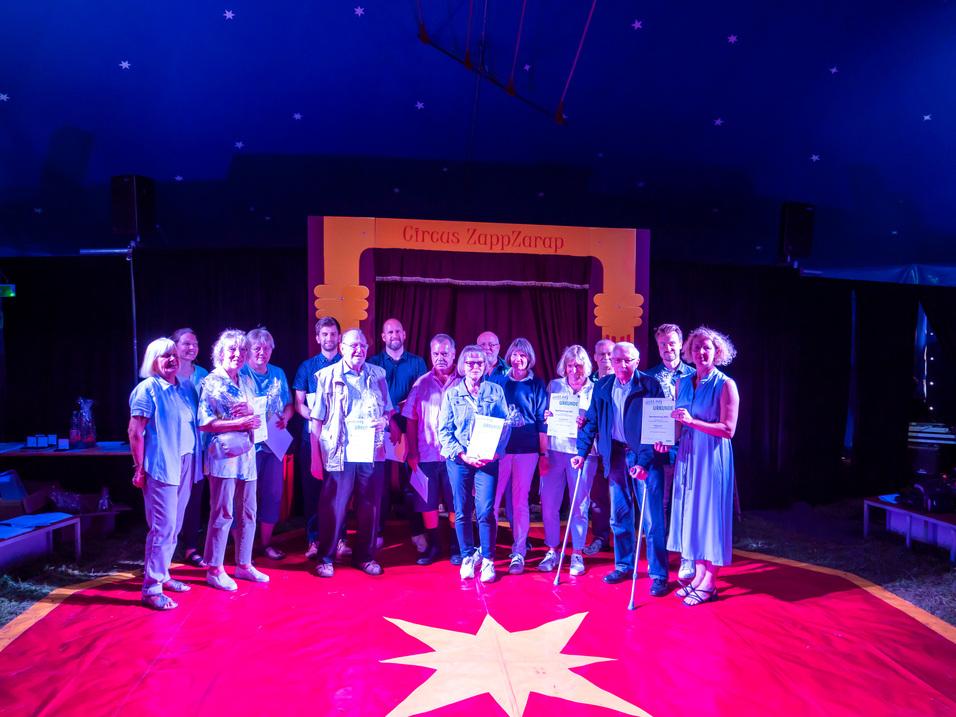 Gruppe von Menschen im Zirkuszelt hält Urkunden, vor rotem Teppich mit goldener Sternform und bunter Dekoration.