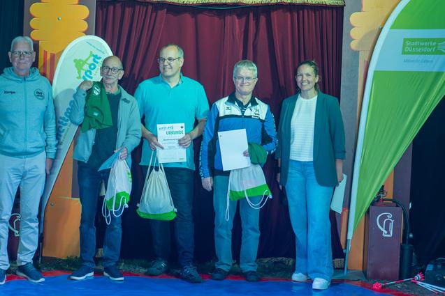 Fünf Personen stehen auf einer Bühne vor einem roten Vorhang, halten Preise und Urkunden in der Hand.