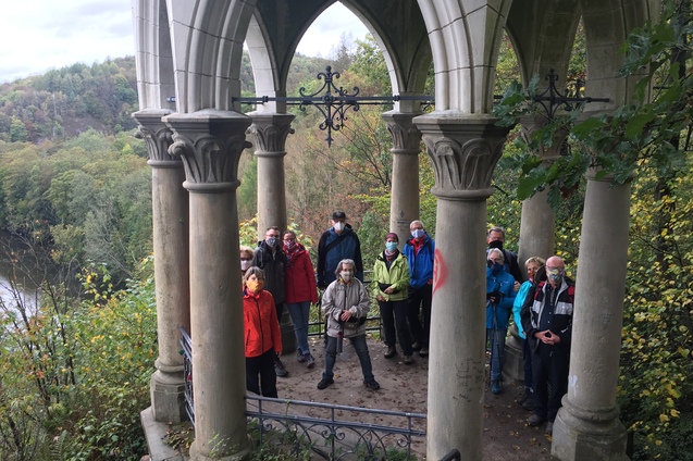 Gruppe von Menschen in Wanderbekleidung steht unter einem gotischen Pavillon mit Säulen, umgeben von Waldlandschaft.