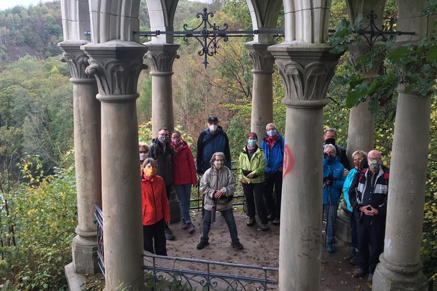 Gruppe von Menschen in Wanderbekleidung steht unter einem gotischen Pavillon mit S&auml;ulen, umgeben von Waldlandschaft.