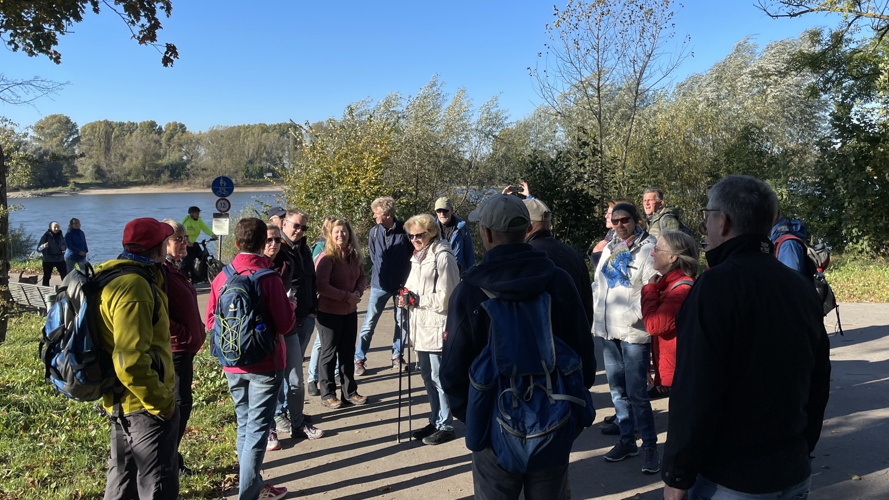 Gruppe von Wanderern in bunter Kleidung versammelt sich am Flussufer unter blauem Himmel.