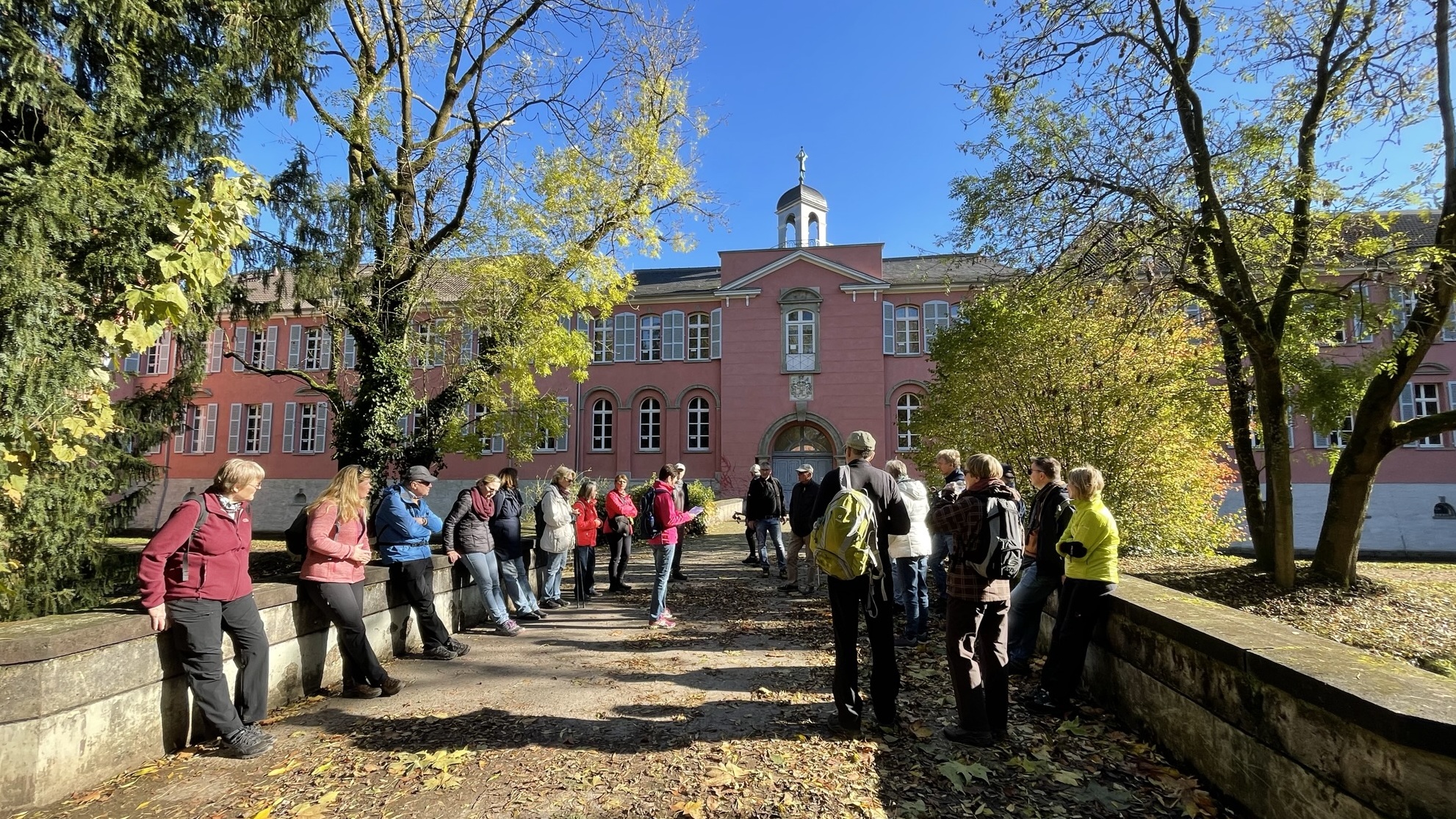 Gruppe von Menschen steht auf einem Weg mit Laub, vor einem rosa Gebäude unter blauem Himmel.