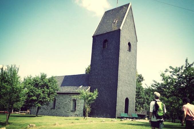 Zwei Personen gehen auf einem Weg auf eine graue Kirche mit einem schmalen Turm in einer grünen Landschaft zu.