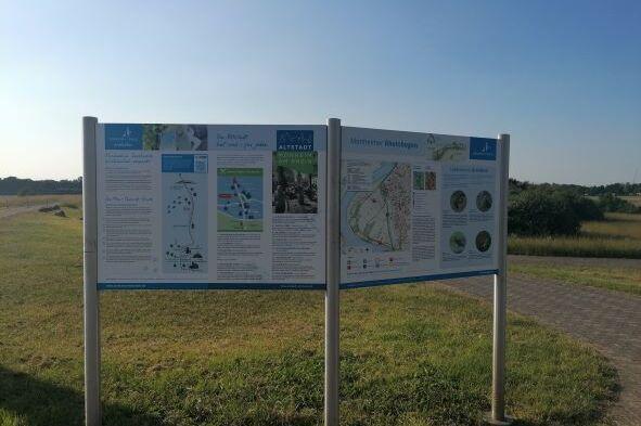 Informationstafel mit Karten und Texten über die Umgebung, auf einem Weg in der Natur aufgestellt.
