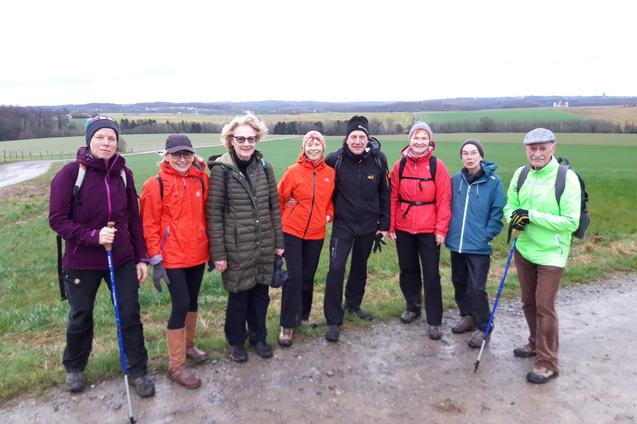Gruppe von acht Wanderern in Umhang und Stiefeln, stehen auf einem Weg mit grünen Feldern im Hintergrund.